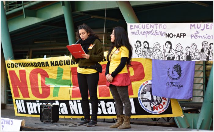 Two women in front of rally, flanked by related banners. One holding a microphone speaking to crowd.